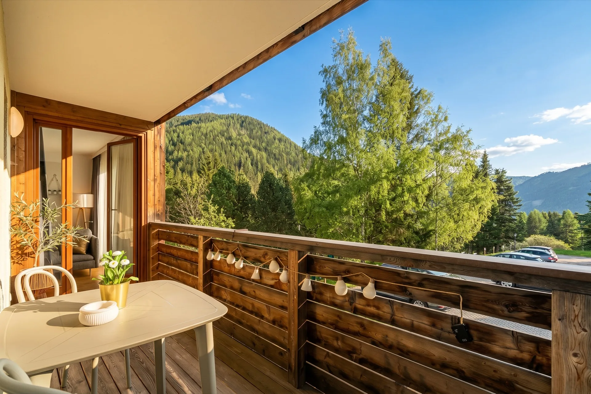 Holzbalkon mit weißem Bistrotisch, Lichterkette und warmem Abendlicht, Blick in das gemütliche Wohnzimmer und auf grüne Berglandschaft beim Rosie Design Chalet Brunnach Bad Kleinkirchheim – Balkonfotografie Ferienwohnung – Mathias Prägant Photography
