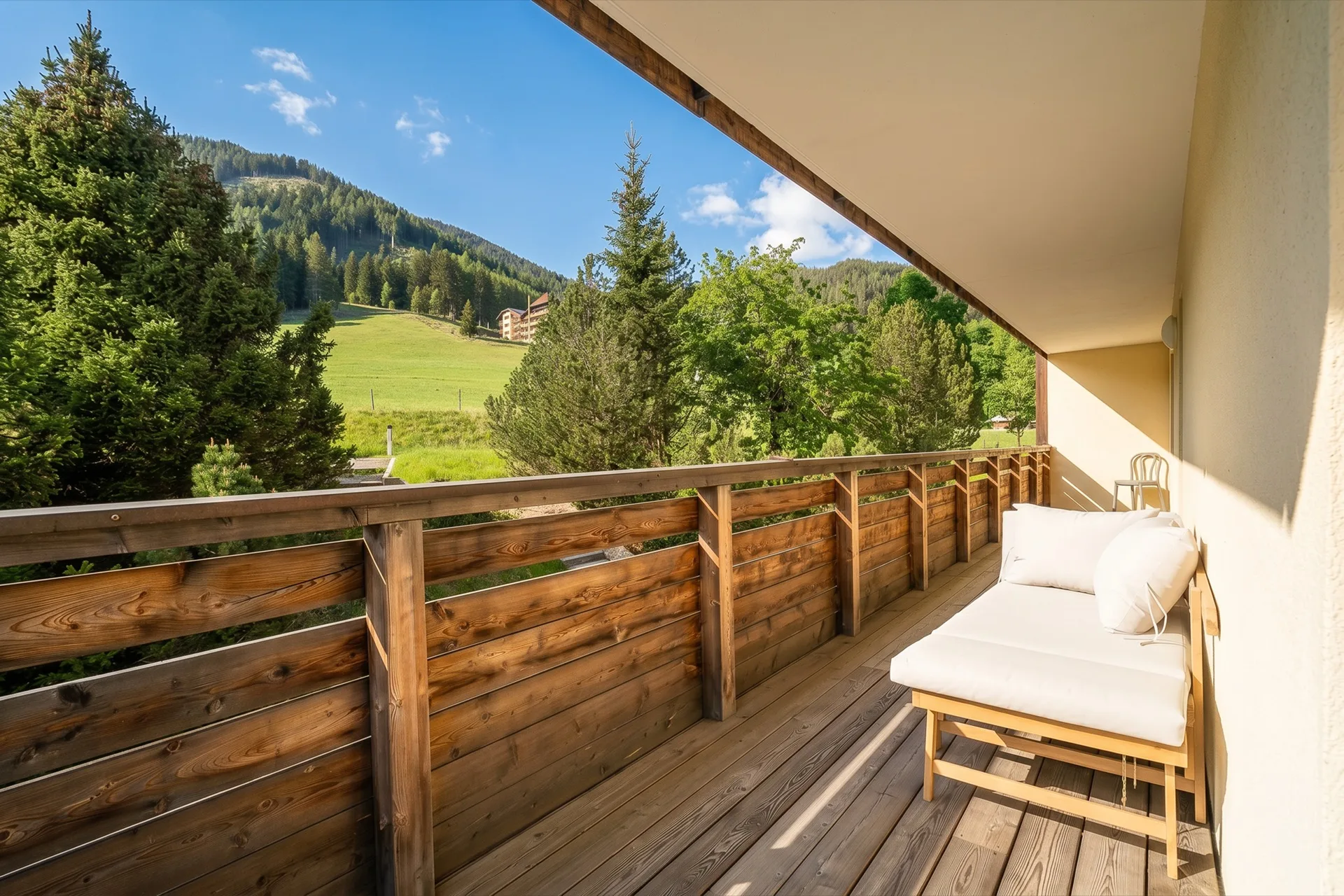 Sonniger Holzbalkon mit weißem Daybed und freiem Blick auf saftige Alpenwiesen, Bergwald und blauen Himmel beim Rosie Design Chalet Brunnach Bad Kleinkirchheim – Balkonfotografie Ferienwohnung Kärnten – Mathias Prägant Photography