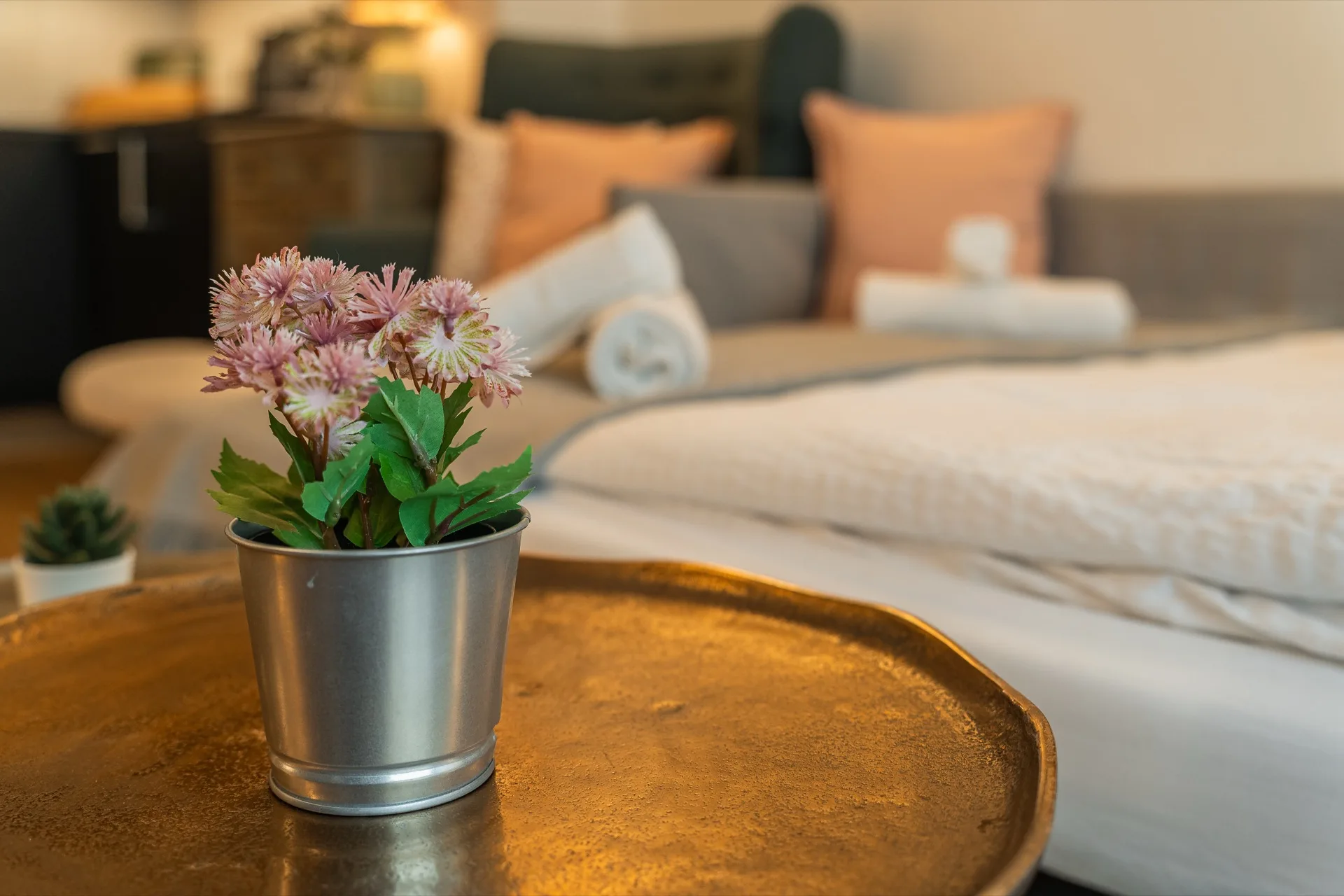 Zartrosa Blumenstrauß in Metalltopf auf goldenem Tablett mit warmem Schlafzimmer-Bokeh im Hintergrund, Rosie Design Chalet Nockalm Bad Kleinkirchheim – Atmosphäre-Detail-Fotografie Ferienwohnung Kärnten – Mathias Prägant Photography