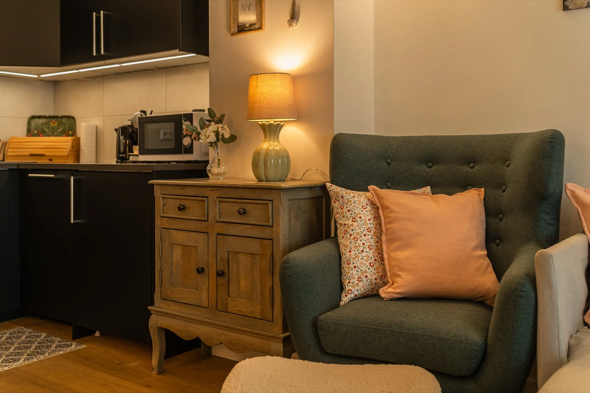 Gemütlicher grüner Ohrensessel mit rosafarbenem Kissen, warmer Tischlampe auf antikem Sideboard und sichtbarer moderner Küche im Hintergrund, Rosie Design Chalet Nockalm Bad Kleinkirchheim – Wohnatmosphäre-Fotografie Ferienwohnung Kärnten – Mathias Prägant Photography