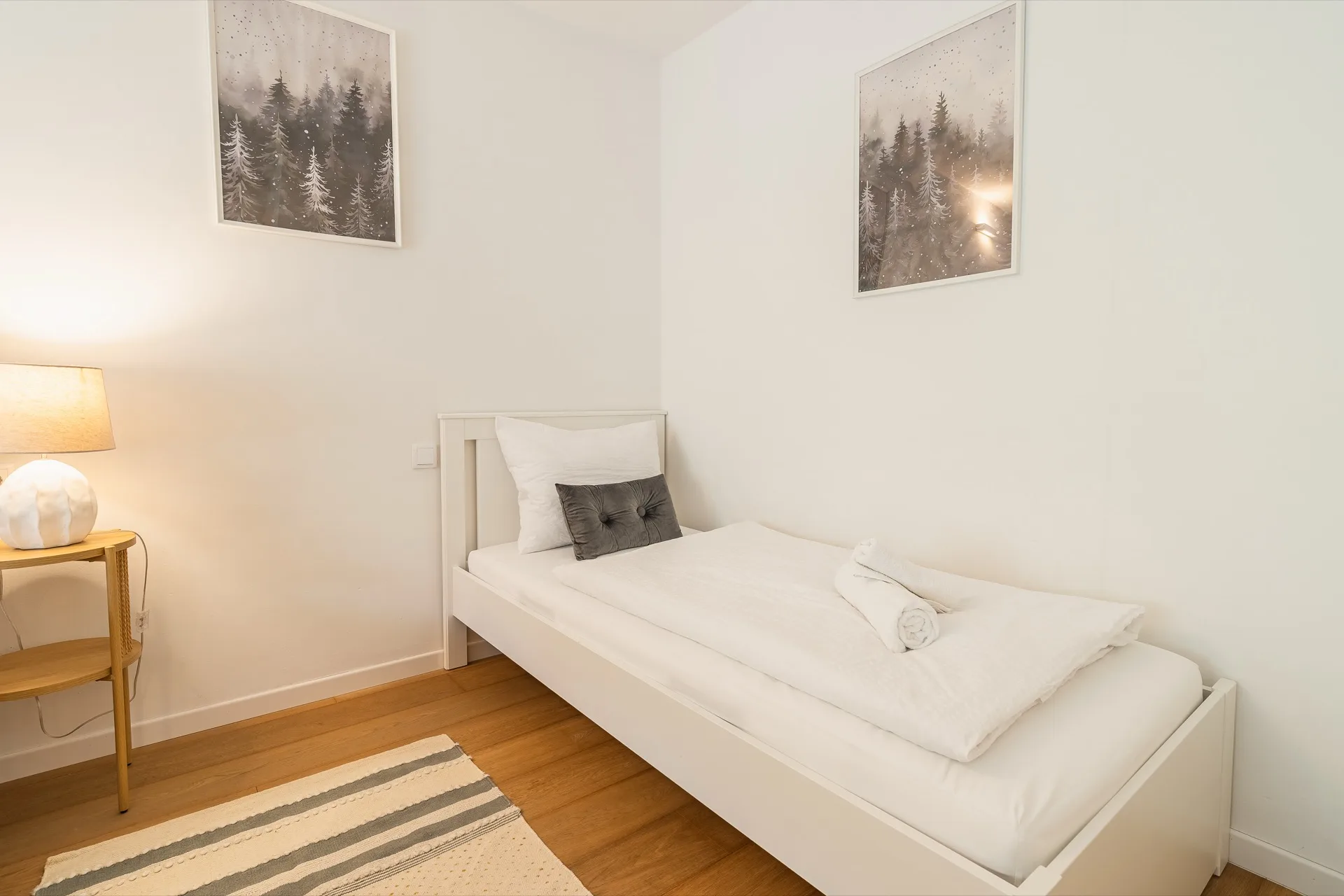 Helles Einzelzimmer mit weißem Bett, Nachttischlampe, Parkett und zwei stimmungsvollen Waldmotiv-Kunstdrucken im Rosie Design Chalet Nockalm Bad Kleinkirchheim – Schlafzimmerfotografie Ferienwohnung Kärnten – Mathias Prägant Photography