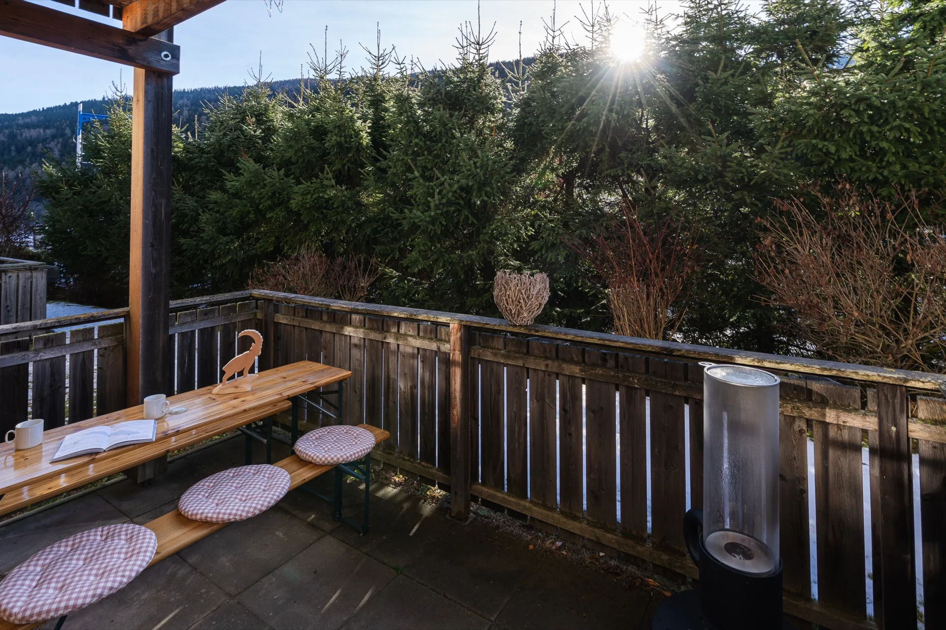 Sonnige Holzterrasse mit Bierzeltgarnitur, Sitzkissen und strahlendem Sonnenstern zwischen alpinen Bäumen der Bergfreunde Bad Kleinkirchheim – Terrassenfotografie Ferienwohnung – Mathias Prägant Photography